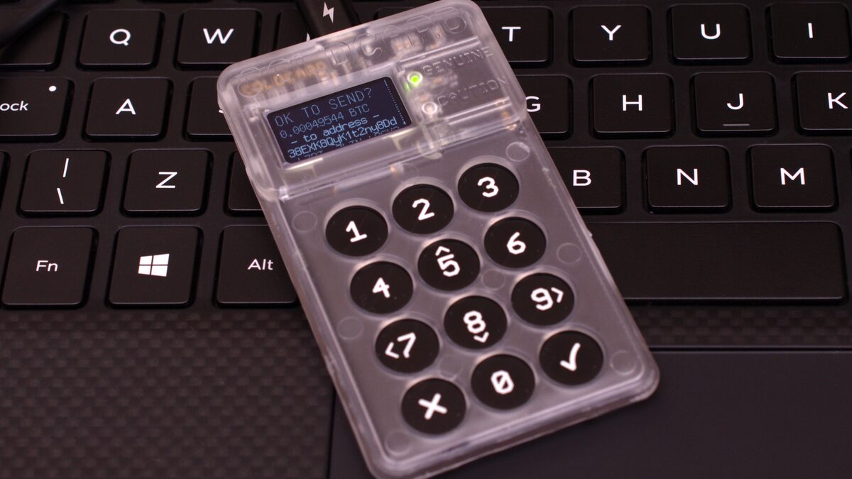 A transparent-cased Coldcard hardware wallet resting on a laptop keyboard, its screen reading 'OK TO SEND?' above a Bitcoin amount and destination address.