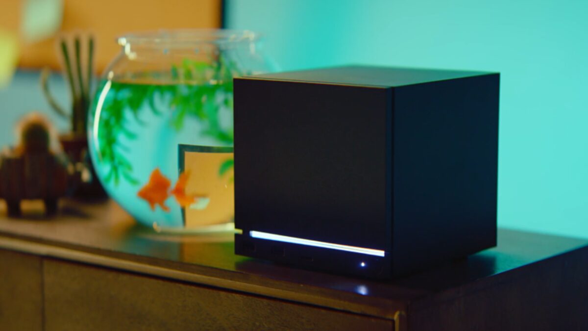 Valve's Steam Machine, a small black cube console, sits on a wooden cabinet beside a fishbowl in a living room.